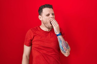 Young hispanic man standing over red background bored yawning tired covering mouth with hand. restless and sleepiness. 