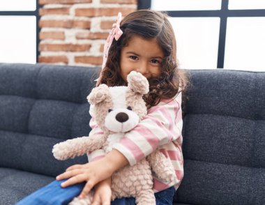 Adorable hispanic girl hugging teddy bear sitting on sofa at home