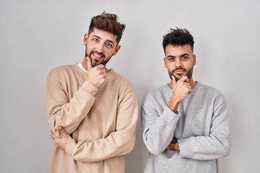 Beyaz arka planda duran genç homoseksüel çift kameraya bakarak kollarını kavuşturup, ellerini çenesine kaldırarak gülümsüyor. Olumlu düşünmek. 