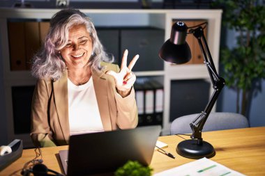 Gri saçlı orta yaşlı bir kadın gece geç saatlerde bilgisayar bilgisayarı kullanarak çalışırken üç numaralı parmaklarını gösterip mutlu ve kendinden emin bir şekilde gülümsüyor.. 