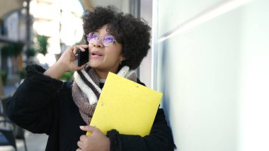 Genç Afrikalı Amerikalı kadın gülümsüyor. Elinde klasörle caddede telefonla konuşuyor.
