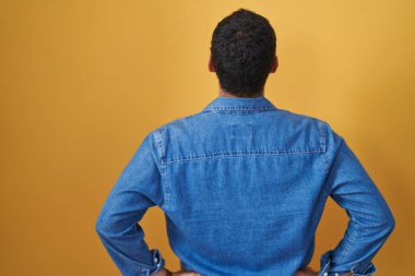 Hispanic man standing over yellow background standing backwards looking away with arms on body 