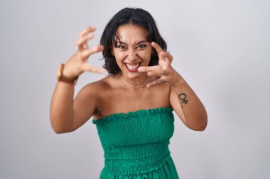 Young hispanic woman standing over isolated background shouting frustrated with rage, hands trying to strangle, yelling mad 