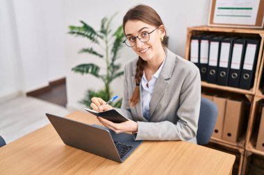 Young caucasian woman business worker using laptop writing on notebook at office
