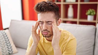 Evdeki kanepede oturan Latin asıllı genç adam baş ağrısı çekiyor.