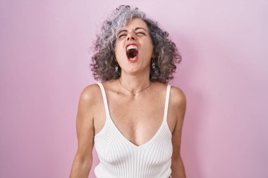Pembe arka planda gri saçlı orta yaşlı bir kadın öfkeli ve öfkeli çığlıklar atıyor, öfkeyle bağırıyor. öfke ve agresif konsept. 