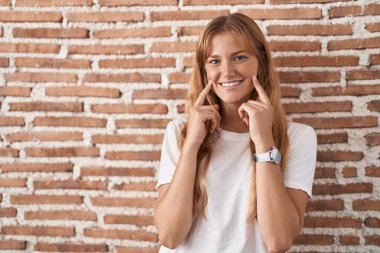 Kafkasyalı genç bir kadın tuğla duvarın üzerinde duruyor ağzı açık gülümsüyor, parmakları işaret ediyor ve neşeli bir gülümsemeye zorluyor. 