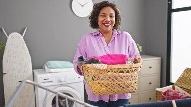 Young beautiful latin woman smiling confident holding basket with clothes at laundry room