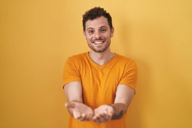 Sarı arka planda duran, avuçlarını birbirine dolamış gülümseyen, el hareketi yapan İspanyol genç bir adam. Bekleme ve koruma 
