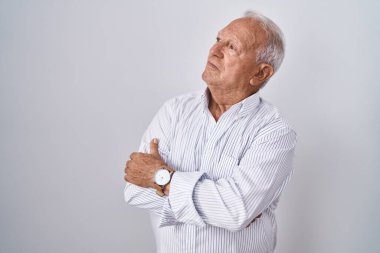 Gri saçlı yaşlı adam izole edilmiş arka planda duruyor. Kollarını kavuşturmuş, ikna olmuş ve kendinden emin bir şekilde yana bakıyor. 