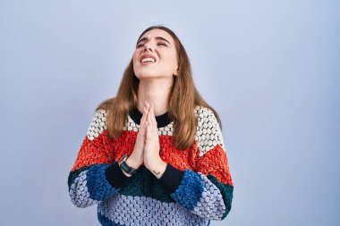 Genç İspanyol kız mavi arka planda duruyor. Elleriyle yalvarıyor ve dua ediyor. Yüzünde umut ifadesi var. Yalvarıyor. 