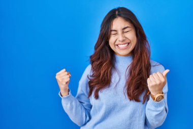 Latin kökenli genç bir kadın mavi arka planda mutlu ve heyecanlı bir şekilde kolları havada, gülümseyen ve başarı için bağıran bir jest yapıyor. kutlama konsepti. 