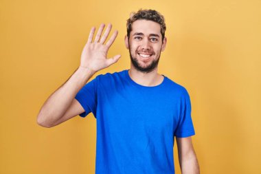 Sarı arka planda duran İspanyol sakallı adam selam veriyor mutlu ve gülümsüyor, dostça karşılama hareketi yapıyor. 