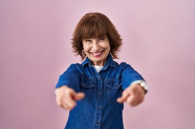 Middle age woman standing over pink background pointing to you and the camera with fingers, smiling positive and cheerful 