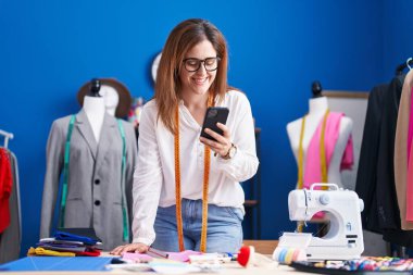 Young woman tailor smiling confident using smartphone at sewing studio