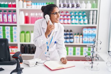 Young beautiful hispanic woman pharmacist talking on telephone writing on notebook at pharmacy