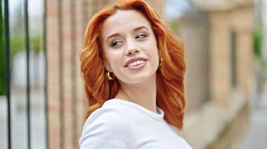 Genç kızıl saçlı kadın gülümsüyor. Kendinden emin bir şekilde caddenin kenarına bakıyor.