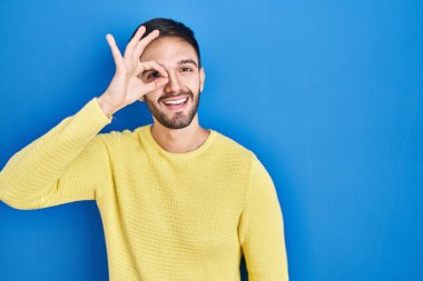Hispanic man standing over blue background doing ok gesture with hand smiling, eye looking through fingers with happy face. 