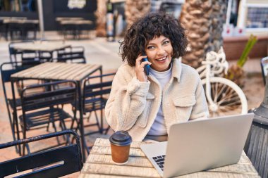 Genç, güzel, İspanyol bir kadın kahve dükkanının terasında dizüstü bilgisayar kullanarak akıllı telefondan konuşuyor.