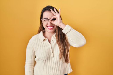 Young hispanic woman standing over yellow background doing ok gesture with hand smiling, eye looking through fingers with happy face. 