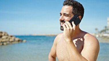 Genç İspanyol turist akıllı telefondan konuşuyor plajda üstsüz duruyor.