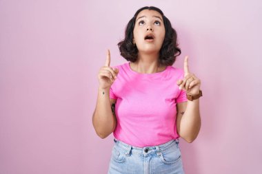 Pembe arka planda duran genç İspanyol kadın şaşırmış ve şaşırmış bir şekilde yukarı bakıyor ve parmaklarıyla işaret ediyor ve kollarını kaldırıyor.. 