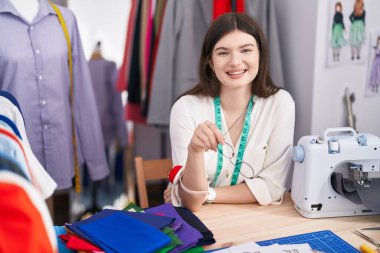 Young caucasian woman tailor smiling confident sitting on table at tailor shop