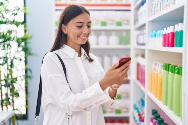 Genç ve güzel İspanyol kadın müşteriler eczanede akıllı telefon kullanarak gülümsüyor.