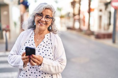 Sokakta akıllı telefon kullanan orta yaşlı bir kadın gülümsüyor.