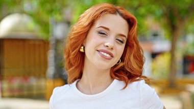 Parkta kendinden emin gülümseyen genç kızıl saçlı kadın.