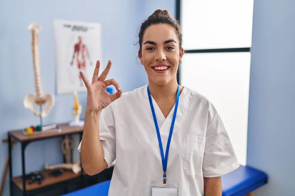 リハビリテーションクリニックで働く若いヒスパニック系女性が手や指でOkサインをして積極的に笑顔 成功した表現 — ストック写真