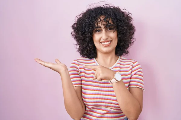 年轻的中东女人站在粉色的背景上 一边手牵着手 一边用手指指着摄像机 一边惊讶地微笑着 — 图库照片