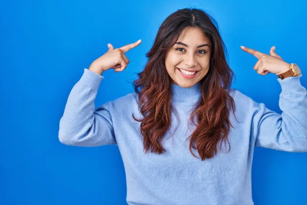 西班牙裔年轻女子站在蓝色的背景上微笑着 用双手指指着头 很好的主意或想法 好的记忆力 — 图库照片