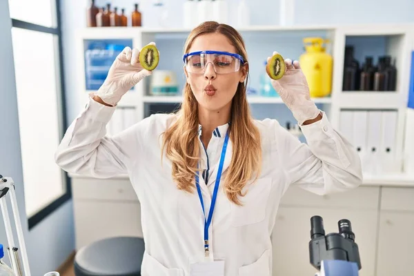 Young blonde scientist woman working with food at laboratory making fish face with mouth and squinting eyes, crazy and comical.