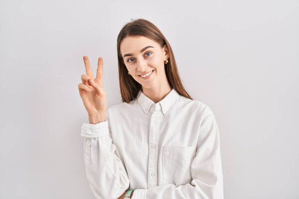 Молодая белая женщина, стоящая на изолированном фоне, улыбаясь, смотрит в камеру, показывая пальцы, делающие знак победы. Номер два. 