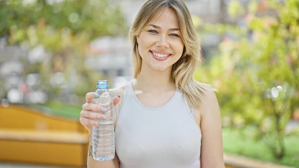 年轻的金发女子笑着自信地在公园里拿着一瓶水 — 图库照片