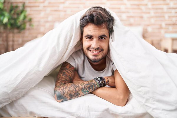 Young hispanic man lying on bed covering with bedsheet at bedroom