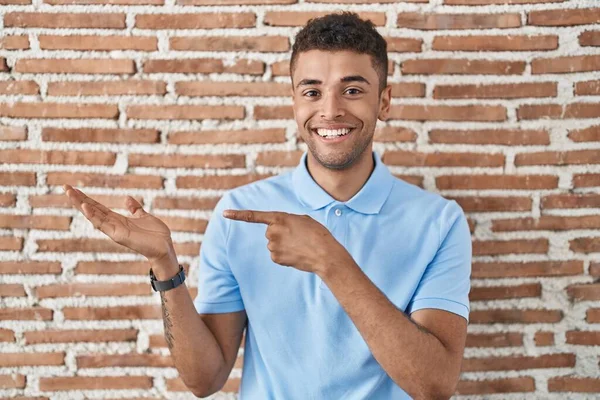 手で提示し 指で指している間 レンガの壁に立っているブラジルの若い男は驚きとカメラに笑顔 — ストック写真
