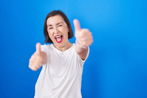 青い背景の上に立つ中年のパニック女性は 手で積極的なジェスチャーを行うことを承認します 親指アップ笑顔と成功のために幸せ 勝者のジェスチャー — ストック写真
