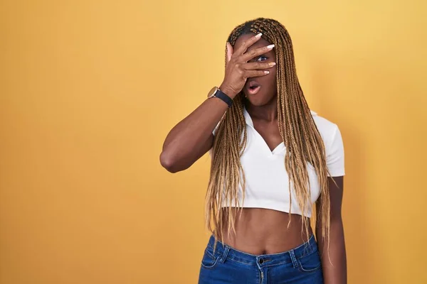 Африканская Американка Плетеными Волосами Стоящая Жёлтом Фоне Подглядывая Шокирующее Лицо — стоковое фото