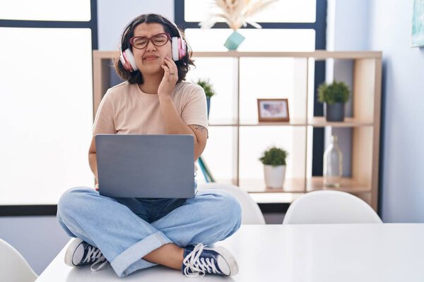 Young hispanic woman using laptop sitting on the table wearing headphones touching mouth with hand with painful expression because of toothache or dental illness on teeth. dentist 
