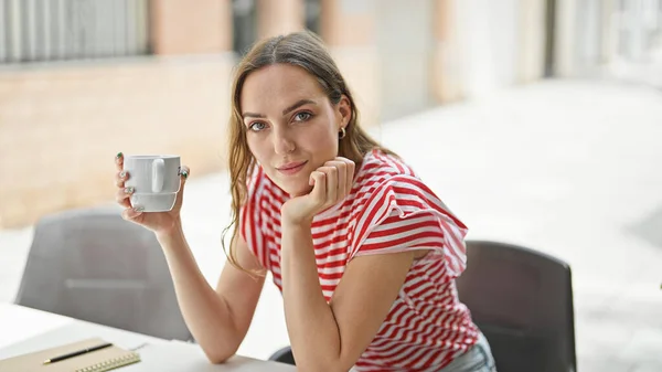 Young blonde woman drinking cup of coffee sitting on table at coffee shop terrace