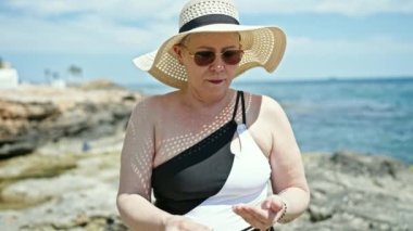 Orta yaşlı, gri saçlı, mayo giyen ve yaz şapkalı bir turist kumsalda koluna güneş kremi sürüyor.