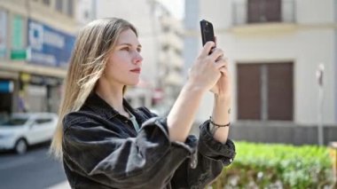 Sokakta gülümseyen akıllı telefonun yanında video kaydeden genç sarışın kadın.