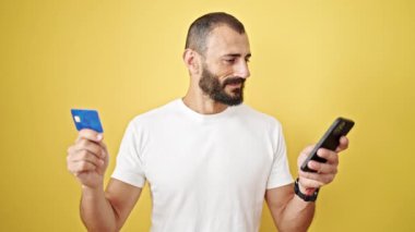 Akıllı telefon ve kredi kartıyla alışveriş yapan İspanyol genç adam izole edilmiş sarı arka planda kutlama yapıyor.