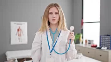 Klinikte steteskop tutan genç sarışın doktor gülümsüyor.