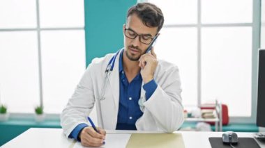 Young hispanic man doctor talking on smartphone writing medical report at clinic