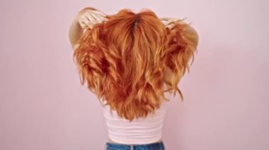 Genç kızıl saçlı kadın pembe arka planda saçlarına dokunuyor.