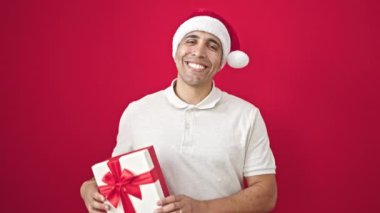 Genç İspanyol adam gülümsüyor. Kendine güveni tam. Noel hediyesini izole edilmiş kırmızı arka planda tutuyor.