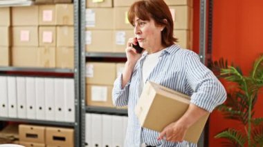 Orta yaşlı kadın ecommerce iş çalışanı elinde paketle ofiste akıllı telefondan konuşuyor.